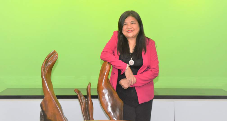 May Yap standing with wooden sculptures against a green backdrop