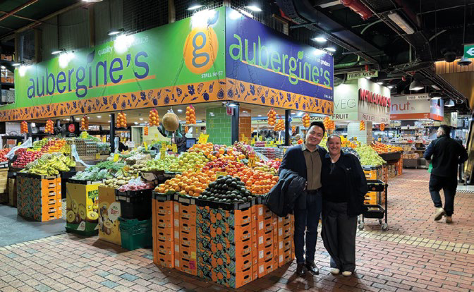 Two people posing at colorful fruit and vegetable market stall