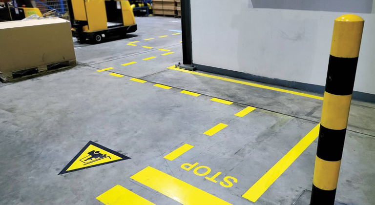 Warehouse floor marked with yellow safety lines, a pedestrian crossing symbol, and a stop sign near a black and yellow bollard